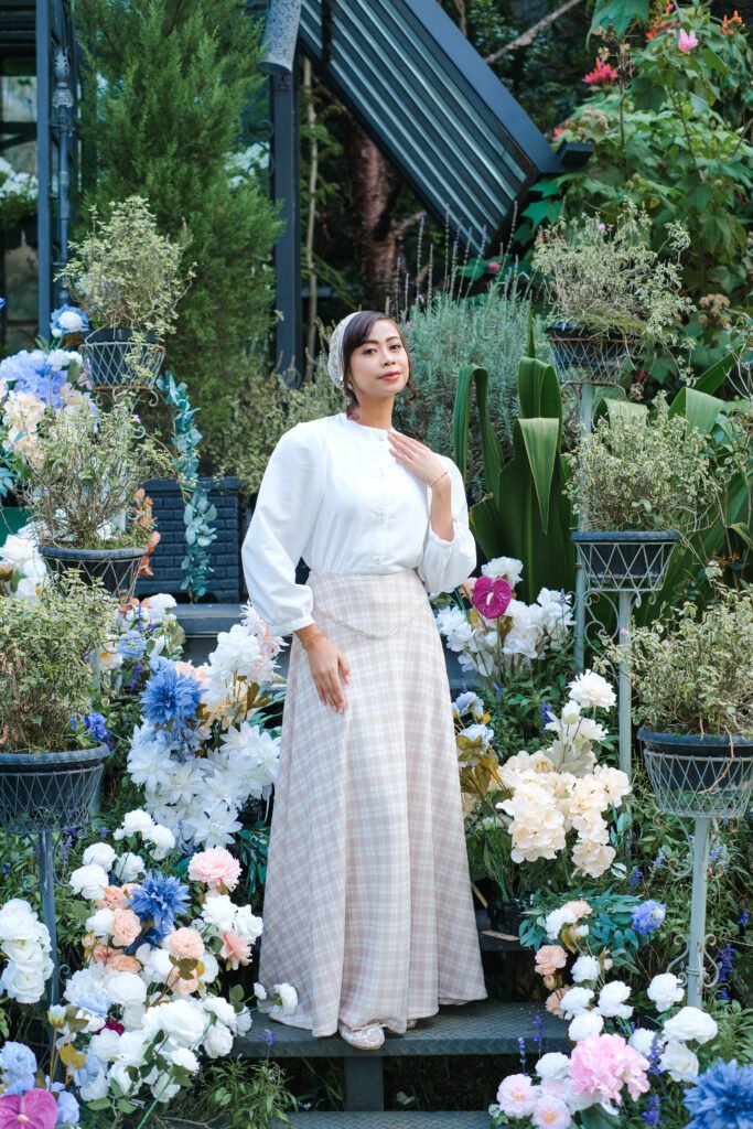 Wanita berdiri di tangga berbunga mengenakan blus putih dan rok kotak-kotak beige.