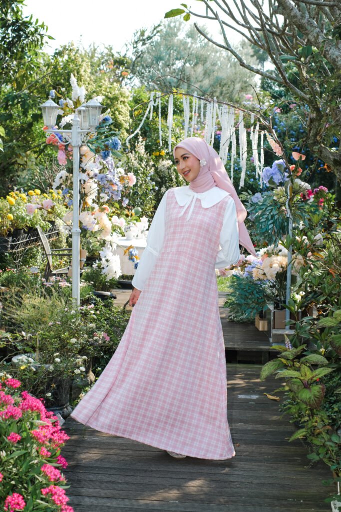 Model muslimah mengenakan overall dress kotak-kotak pink dan kemeja putih di taman.