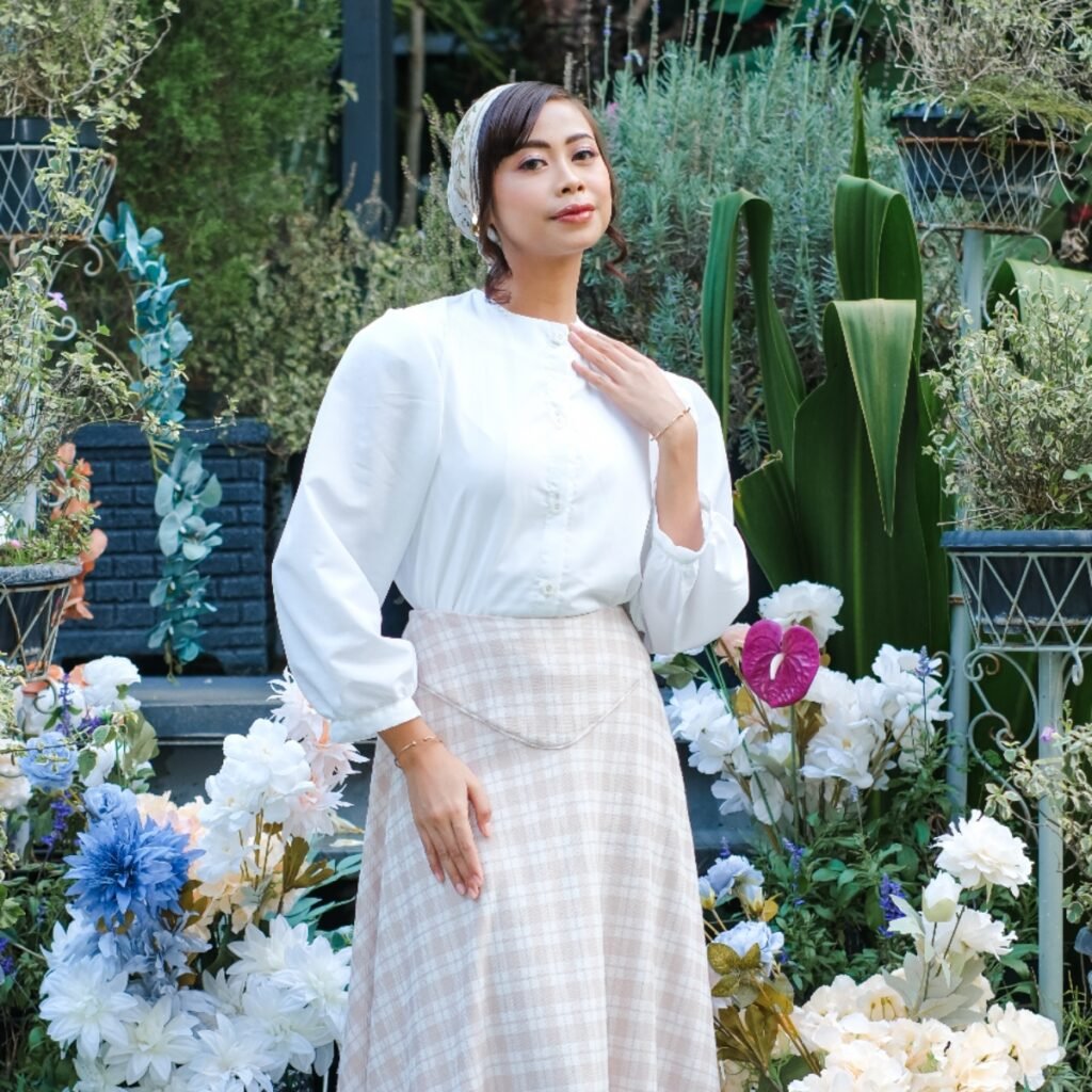 Wanita berdiri di tangga berbunga mengenakan blus putih dan rok kotak-kotak beige.