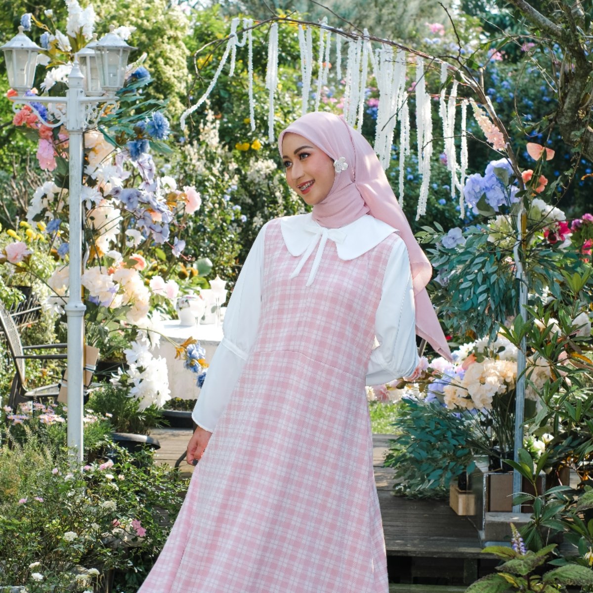 Model muslimah mengenakan overall dress kotak-kotak pink dan kemeja putih di taman.